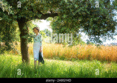 Donna in 40's vestiti sotto un albero di olivo nella campagna abruzzese con un campo di grano in background Foto Stock