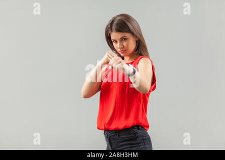 Ritratto di grave bella bruna giovane donna in camicia rossa in piedi con la boxe pugni e guardando la telecamera, pronto per l'attacco o difesa. interni, Foto Stock