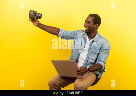 Ritratto di Allegro fotografo amatoriale con sorriso toothy in denim shirt tenendo selfie foto digitali con fotocamera DSLR e azienda portatile, di video live Foto Stock