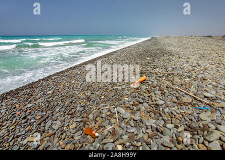 Vari rifiuti di plastica sulla selvaggia incontaminata di ciottoli neri spiaggia di pietra, uno splendido mare in background. Foto Stock
