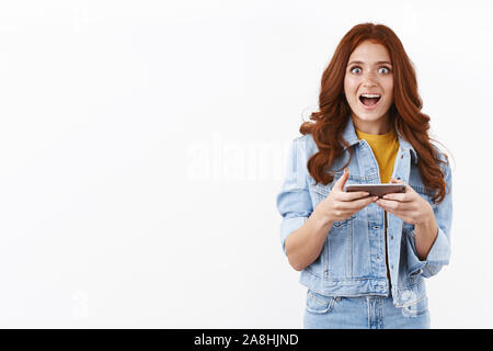 Emozionato allegro redhead donna in camicia di denim, tenere lo smartphone orizzontalmente, affannosa divertito e lo sguardo attonito fotocamera, vincendo il livello hard in mobile Foto Stock