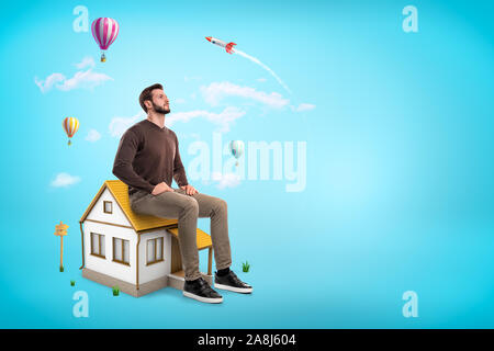 Giant giovane uomo seduto sul tetto della casa piccola come su una sedia, guardando in alto nel cielo blu con copia spazio e sognare. Foto Stock