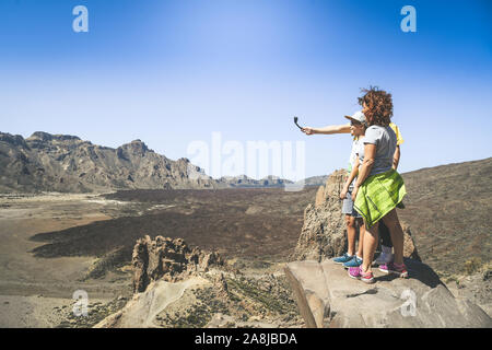 Mamma e figli godendo all'aperto in piedi su una scogliera di montagna che si affaccia sul Tenerife Teide National Park. Spensierato giovane famiglia tenendo selfie con act Foto Stock