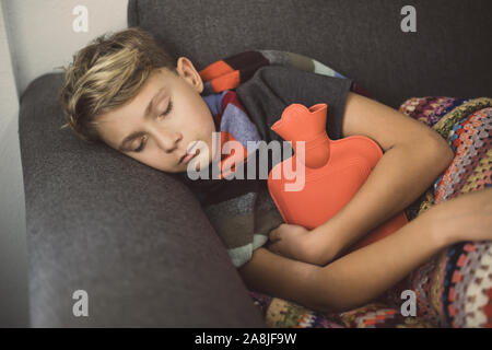 Ragazzo malato a letto con coperta di lana e bottiglie di acqua calda. Triste teen con l'influenza si appoggia a casa da solo in una fredda giornata invernale. Bambino con infettare stagionali Foto Stock