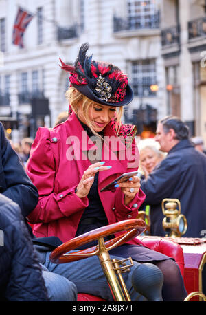 Signora giovane in vintage abbigliamento vittoriano seduti su auto d'epoca, guardando il suo telefono cellulare, creando un anacronismo Foto Stock