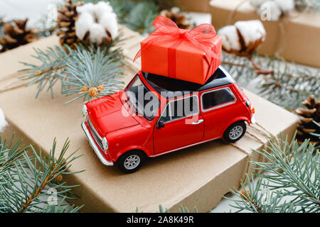 Rosso auto giocattolo con un Natale confezione regalo rossa sul tetto. Sfondo di natale con cotone, Abete rami, abete coni. Vacanze di Natale, celebrazione Foto Stock