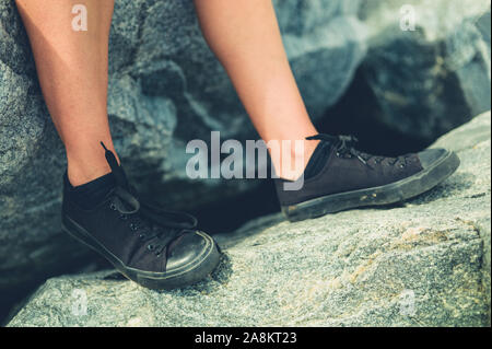 I piedi di una giovane donna che indossa scarpe nere seduto su una roccia Foto Stock