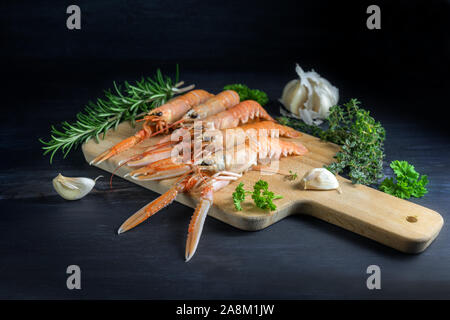 Scampi freschi, chiamato anche Scampo langoustine o su una scheda di cucina, anche aglio ed erbe aromatiche, gli ingredienti per un festoso pasto a base di pesce al buio su un g Foto Stock