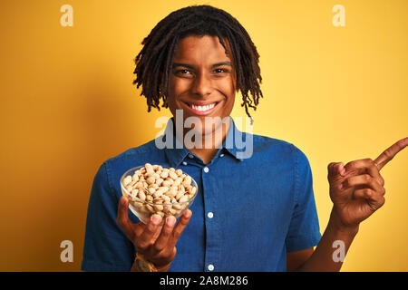 Giovani afro-americano con dreadlocks di mangiare i pistacchi isolato su sfondo giallo molto felice rivolto con la mano e le dita per lato Foto Stock