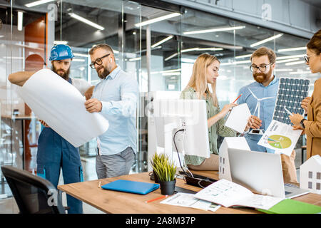 Un gruppo di giovani ingegneri che lavorano su un progetto di energia alternativa, senior caposquadra e lavoratore in piedi con blueprint in ufficio Foto Stock