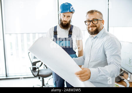 Ritratto di un senior caposquadra e operaio in tuta a lavorare sul progetto con blueprint in ufficio Foto Stock