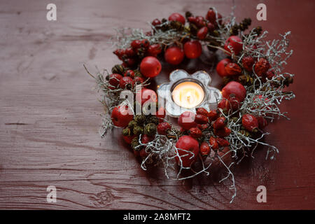 Ghirlanda di naturale con rosa canina e piccole mele intorno ad una candela che brucia in un rosso tavolo in legno, decorazioni stagionali in autunno, Avvento e Natale Foto Stock