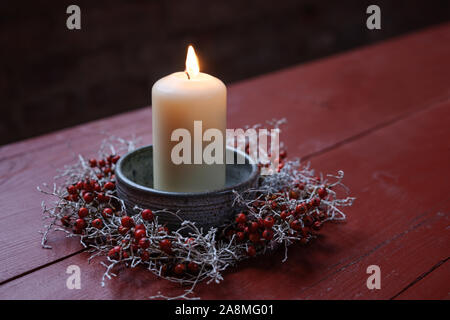 Candela che brucia in una ciotola decorata con una corona piccola dalla rosa canina e argento cuscino sulla boccola in rosso di un tavolo di legno, disposizione invernale per l Avvento, ch Foto Stock