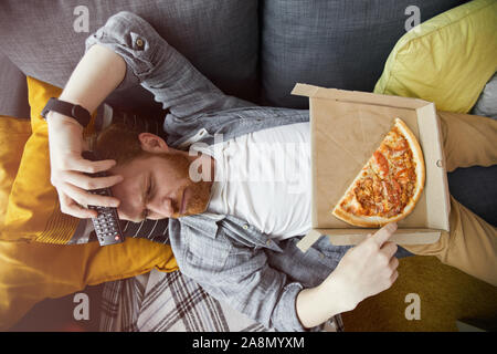 Vista sopra il ritratto di uomo barbuto sdraiato sul lettino e mangiare la pizza mentre si guarda la TV di casa, spazio di copia Foto Stock