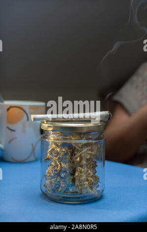 Fumare un giunto di marijuana con una tazza di caffè a casa. Riutilizzare il vasetto di vetro pieno di boccioli di cannabis con un erbaccia sigaretta, vicino. Copia superiore dello spazio. Foto Stock