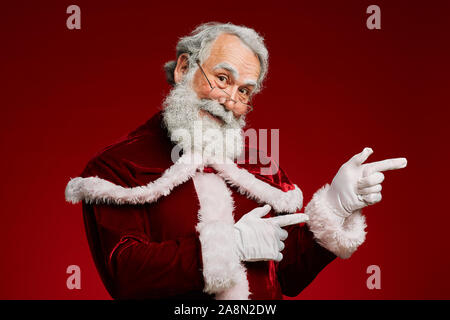 Vita ritratto di sorridere Santa Claus guardando la fotocamera e puntando a lato mentre in piedi contro lo sfondo di colore rosso, spazio di copia Foto Stock