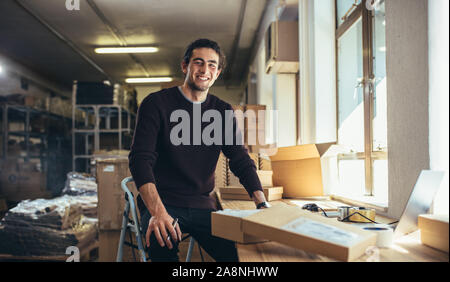 Online di successo titolare di azienda seduto alla sua scrivania. Giovane Uomo con laptop e scatola di spedizione sulla scrivania, guardando la fotocamera e sorridente. Foto Stock