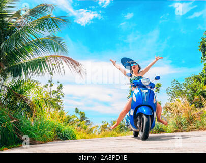 Crazy funny donna con capelli volanti in sella a una moto su un cielo blu e verde tropici sfondo. Ragazza giovane con i capelli scuri in occhiali da sole su un blu Foto Stock
