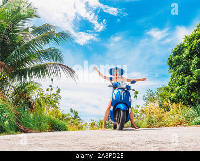 Crazy funny donna con capelli volanti in sella a una moto su un cielo blu e verde tropici sfondo. Ragazza giovane con i capelli scuri in occhiali da sole su un blu Foto Stock