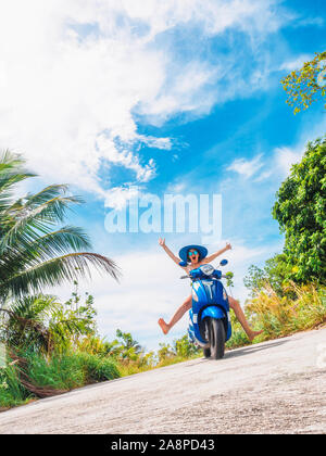 Crazy funny donna con capelli volanti in sella a una moto su un cielo blu e verde tropici sfondo. Bizzarro giovane ragazza con i capelli scuri in occhiali da sole su un Foto Stock