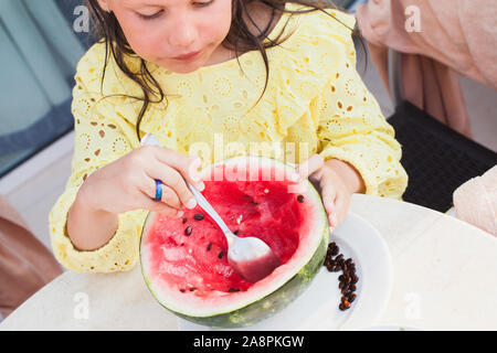 Bambina in abito giallo mangia anguria con un cucchiaio Foto Stock