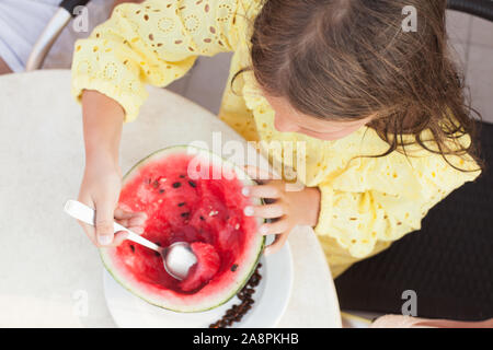 Piccola ragazza caucasica mangia anguria a fette con un cucchiaio, vista dall'alto Foto Stock