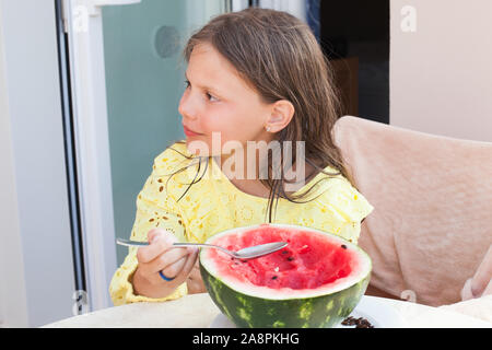 Piccola ragazza caucasica mangia anguria a fette con un cucchiaio, dehor estivo ritratto Foto Stock