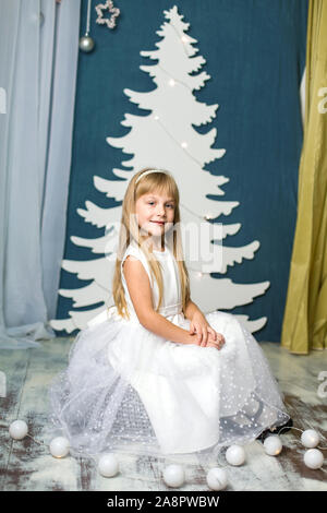 Una piccola ragazza in un abito bianco siede su un puff contro uno sfondo di una artificiale White Christmas tree decor da cartone, e una ghirlanda di fiori si trova accanto Foto Stock