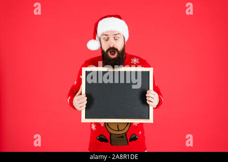 Incredibile. Annuncio d'inverno. Lavagna per informazioni. Man tenere lavagna vuota copia spazio. Guy babbo natale sfondo rosso. Evento invernale. Vacanze invernali. Santa Claus hat con la pubblicità. Foto Stock