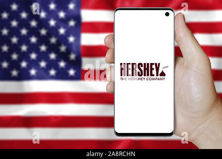 Il logo della società pubblica la Hershey Company visualizzata su uno smartphone. Bandiera degli Stati Uniti sullo sfondo. Credito: PIXDUCE Foto Stock