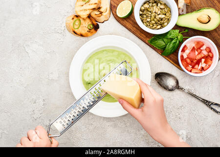 Vista ritagliata dell'uomo grattugiare il formaggio in verde zuppa cremosa vicino al tagliere di legno con ingredienti freschi Foto Stock