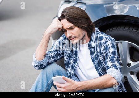 Turbare l uomo in abiti casual tenendo lo smartphone in mano e seduta vicino rotta automatica con tronco aperto, assicurazione auto concept Foto Stock