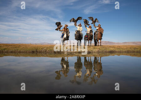 Un gruppo di tradizionale kazako cacciatori eagle mantenendo le loro aquile reali a cavallo sul bordo del fiume con le loro riflessioni. Ulgii, Mongolia. Foto Stock