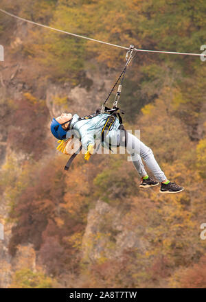 Una ragazza adolescente vola su un zipline contro lo sfondo delle montagne coperte di bosco d'autunno. Foto Stock