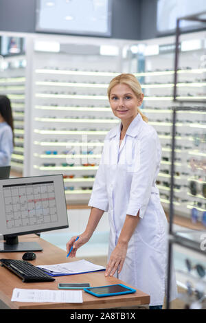 Donna bionda che lavora nel negozio ottica in piedi vicino al tavolo di lavoro Foto Stock