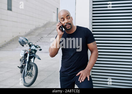 Fiducioso bello mucular uomo in piedi alla parete edilizia e chiamare amico dopo equitazione sulla motocicletta Foto Stock