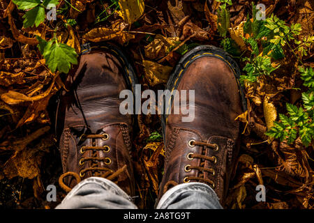 Camminare in un autunno foglia-foresta coperta in stivali in pelle con cuciture gialle e jeans denim da una prospettiva in prima persona che guarda il gro Foto Stock