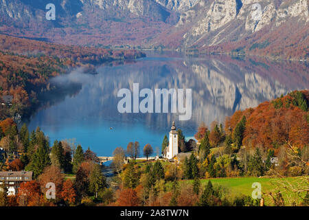 Magnifica vista sul lago di Bohinj e la chiesa di San Giovanni Battista in i colori dell'autunno in una mattina di sole in Slovenia. Viaggi, Turismo e bellezza Foto Stock