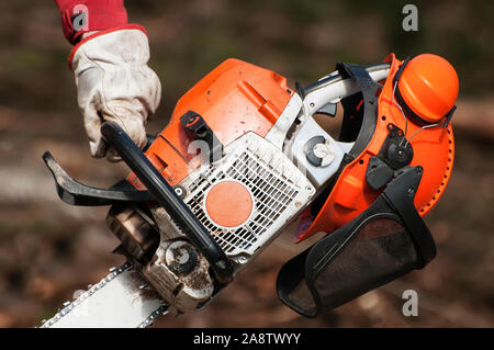 Primo piano della forestazione della mano sinistra tenendo un chainsaw e casco protettivo con cuffie antirumore in appoggio su di esso Foto Stock