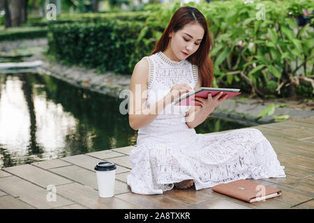 Giovane vietnamita studente di college seduta nel parco con tazza di caffè e mediante applicazione sul computer tablet Foto Stock