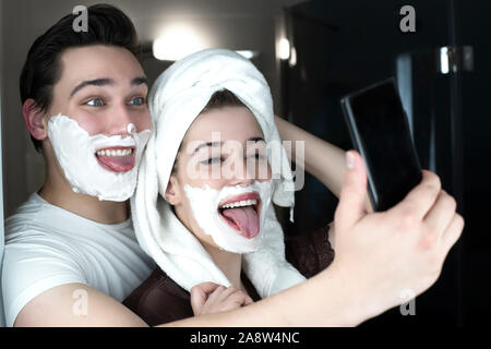 Funny giovane uomo bello e bella donna che colpisce una posa per selfie divertirsi sia con schiuma da barba sui loro volti in bagno allegro. Foto Stock