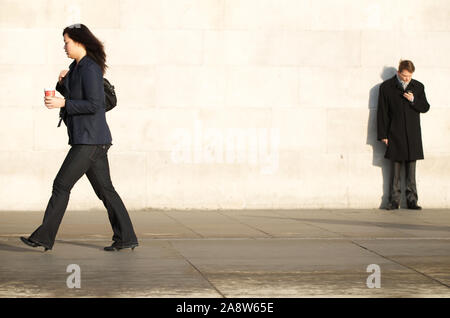 Londra - 17 novembre 2011: una donna che porta una tazza di caffè passa un uomo che sta guardando il suo telefono cellulare in Trafalgar Square. Foto Stock