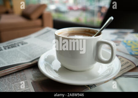 Una tazza piena di caffè in un singolo bicchiere bianco,appoggiata su quotidiani accanto a una finestra esterna con sfondo sfocato. Foto Stock