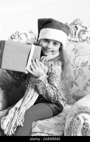 Il tempo di Natale e la sorpresa del concetto. Santas helper nella Red hat si siede sul braccio chic sedia e detiene confezione regalo. Ragazza con bella faccia su sfondo bianco. Adorable kid riceve presenta. Foto Stock