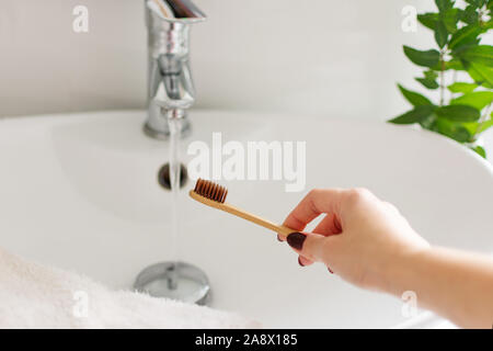 Donna mano utilizzando bio-degradabile toothbrushe bambù in un bagno interno bianco. Pianta verde decor in background. Foto Stock