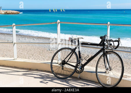 Una bicicletta si siede parcheggiato lungo una rampa per la spiaggia come un gruppo di adattamento di barche a vela sono sfocate in lontananza sulla Riviera Francese a Menton, Francia Foto Stock