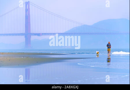 Vista del Golden Gate Bridge con gente e il suo cane che fa jogging lungo la East Beach a San Francisco all'alba. Foto Stock