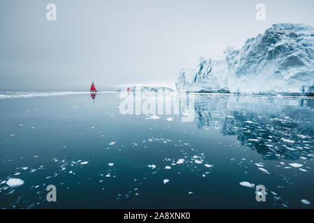 Bella rossa di barca a vela in artico accanto a un enorme iceberg che mostra la scala. Navigando tra iceberg galleggianti nella baia di Disko glacier durante midnigh Foto Stock