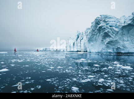 Bella rossa di barca a vela in artico accanto a un enorme iceberg che mostra la scala. Navigando tra iceberg galleggianti nella baia di Disko glacier durante midnigh Foto Stock