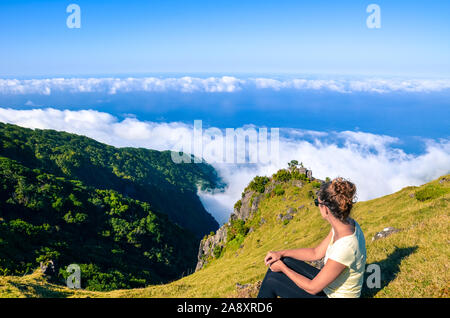 Viaggiatore femmina seduto sulla cima della collina ha una fantastica vista del paesaggio a Madeira, Portogallo. Le colline rocciose e foreste, blu oceano Atlantico in background. Sopra le nuvole. Concetto di viaggio. Foto Stock